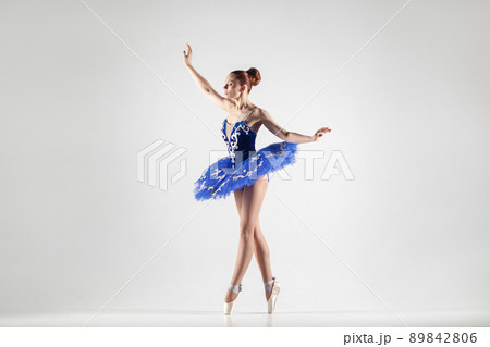 attractive ballerina with bun collected hair wearing blue dress and pointe shoes performing in white studio. indoor, studio shot. attractive ballerina with bun collected hair wearing blue dress and pointe shoes performing in white studio. indoor, studio shot. 89842806