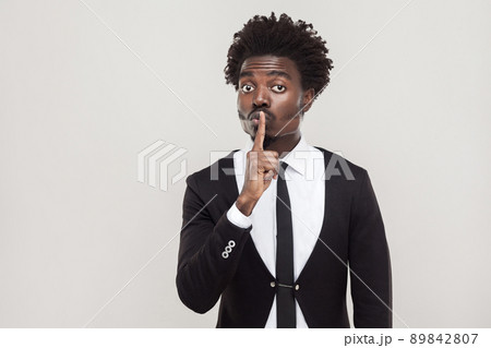 Secret or quiet concept. African businessman showing shh sign at camera. Indoor shot, gray background Secret or quiet concept. African businessman showing shh sign at camera. Indoor shot, gray background 89842807