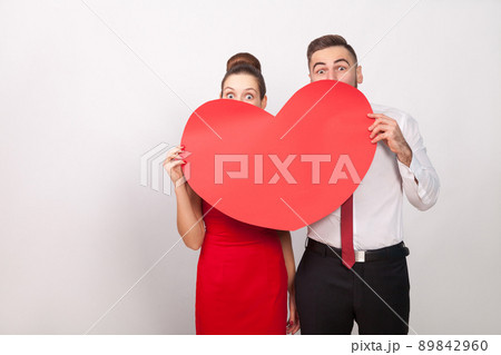 Funny couple hide and seek behind big red heart. Indoor, studio shot, isolated on gray background Funny couple hide and seek behind big red heart. Indoor, studio shot, isolated on gray background 89842960