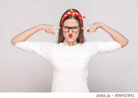 i dont want to hear you. portrait of unsatisfied emotional young woman in white t-shirt with freckles, black glasses, red lips and head band. indoor studio shot, isolated on light gray background. i dont want to hear you. portrait of unsatisfied emotional young woman in white t-shirt with freckles, black glasses, red lips and head band. indoor studio shot, isolated on light gray background. 89842991