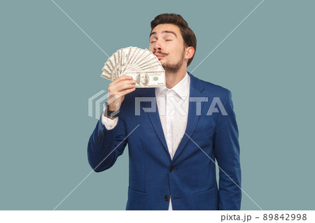 Handsome businessman in blue jacket standing and holding many dollars in hand and smelling and feeling smell of richness with closed eyes. Indoor, studio shot isolated on light blue background. 89842998
