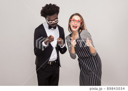 Mixed race couple pointing fingers at camera and smiling. Studio shot, gray background Mixed race couple pointing fingers at camera and smiling. Studio shot, gray background 89843034