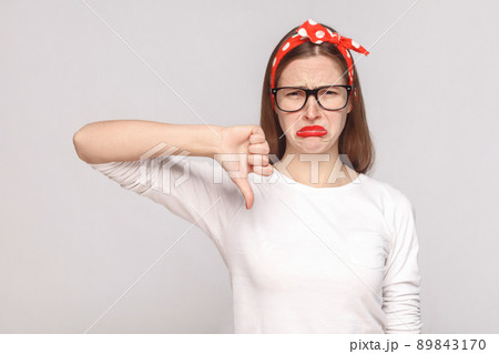 its bad, sad thumbs down of unsatisfied emotional young woman in white t-shirt with freckles, black glasses, red lips and head band looking at camera. studio shot, isolated on light gray background. 89843170