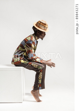Unhappy afro man in traditional national clothes sit on cube. Indoor, isolated on gray background Unhappy afro man in traditional national clothes sit on cube. Indoor, isolated on gray background 89843271