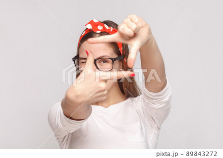choosing composition or focusing portrait of beautiful emotional young woman in white t-shirt with freckles, glasses, red lips and head band. indoor studio shot, isolated on light gray background. 89843272