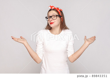 I dont know. i have no idea. portrait of beautiful emotional young woman in white t-shirt with freckles, black glasses, red lips and head band. indoor studio shot, isolated on light gray background. I dont know. i have no idea. portrait of beautiful emotional young woman in white t-shirt with freckles, black glasses, red lips and head band. indoor studio shot, isolated on light gray background. 89843351
