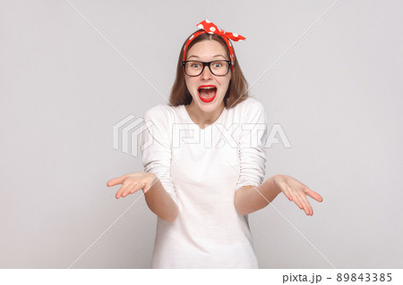 how you did this? it's unbelievable. portrait of beautiful emotional young woman in white t-shirt with freckles, black glasses, red lips and head band. indoor shot, isolated on light gray background. 89843385