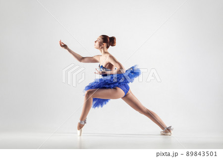 Young beautiful ballerina with bun collected hair wearing blue dress and pointe shoes dancing gracefully isolated on white background. indoor, studio shot. 89843501