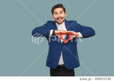 Handsome man in blue jacket and white shirt, standing and holding red heart shape and looking at camera with toothy smile. Indoor, studio shot isolated on light blue background 89843596
