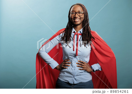 Proud superhero woman with superpower abilities wearing mighty hero cloak while smiling confident at camera. Joyful happy smiling young adult person posing as justice defender on blue background. Proud superhero woman with superpower abilities wearing mighty hero cloak while smiling confident at camera. Joyful happy smiling young adult person posing as justice defender on blue background. 89854261