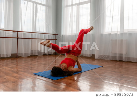 A young woman fitness instructor in red Sportswear Leggings and Top stretching in the gym before her pilates, on a yoga mat near the large window on a sunny day, female fitness yoga routine concept. 89855072