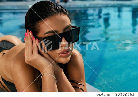 Close up of slim girl in black swimsuit and sunglasses lying on abdomen. Seductive young female relaxing, sunbathing, having vacation. Concept of leisure, summertime and youth. Close up of slim girl in black swimsuit and sunglasses lying on abdomen. Seductive young female relaxing, sunbathing, having vacation. Concept of leisure, summertime and youth. 89863963