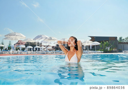 Front view of pretty slim girl swimming in pool, sunbathing, relaxing. Brunette young female with long wet hair standing in water, touching hair with closed eyes. Concept of rest. Front view of pretty slim girl swimming in pool, sunbathing, relaxing. Brunette young female with long wet hair standing in water, touching hair with closed eyes. Concept of rest. 89864036