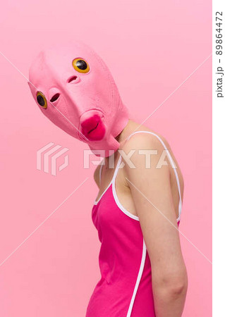 A woman wearing a silicone Halloween mask in the shape of a pink fish with big yellow eyes looks at the camera against a pink background 89864472
