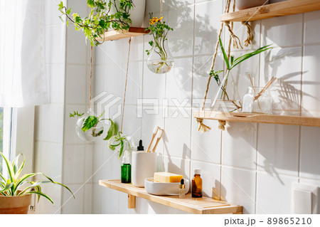 Wooden shelves with cosmetics and toiletries against white tile wall with biophilic and eco friendly design. Hanging glass pots with green plants Wooden shelves with cosmetics and toiletries against white tile wall with biophilic and eco friendly design. Hanging glass pots with green plants 89865210