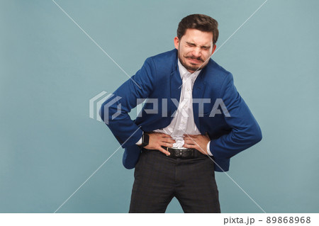 Unhappy man with stomach pain spasm. portrait of handsome bearded businessman in blue suit and white shirt, with smart watch. Indoor studio shot, isolated on light blue background Unhappy man with stomach pain spasm. portrait of handsome bearded businessman in blue suit and white shirt, with smart watch. Indoor studio shot, isolated on light blue background 89868968