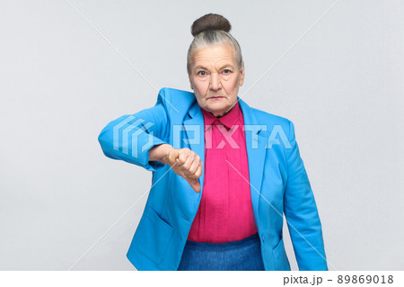 Unhappy woman showing dislike sign. Emotion and feelings, expressive grandmother with light blue suit and pink shirt standing with collected bun gray hair. Studio shot, isolated on gray background Unhappy woman showing dislike sign. Emotion and feelings, expressive grandmother with light blue suit and pink shirt standing with collected bun gray hair. Studio shot, isolated on gray background 89869018