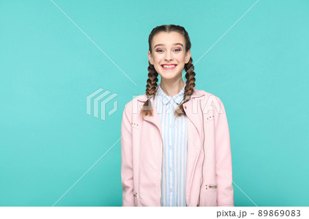 happy toothy smile portrait of beautiful cute girl standing with makeup and brown pigtail hairstyle in striped light blue shirt pink jacket. indoor, studio shot isolated on blue or green background. 89869083