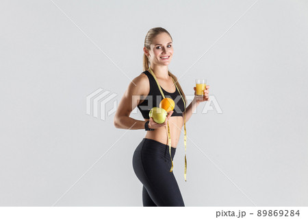 Athletic girl holding fruits and juice, toothy smiling and looking at camera. Indoor shot, gray background 89869284