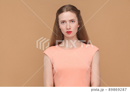 unhappy, sad woman looking at camera. portrait of emotional cute, beautiful woman with makeup and long hair in pink dress. indoor, studio shot, isolated on light brown or beige background. 89869287