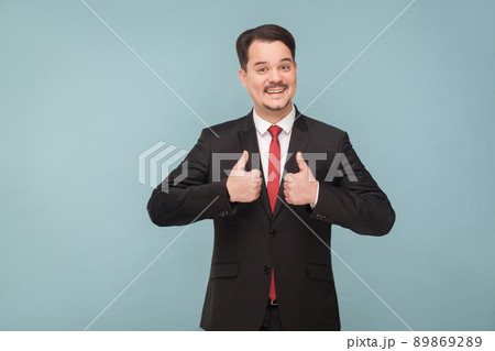 thumbs up handsome businessman in classic black suit showing like sign and looking at camera with toothy smile with mustache, laugh. Front view. Isolated on light blue background looking at camera. 89869289
