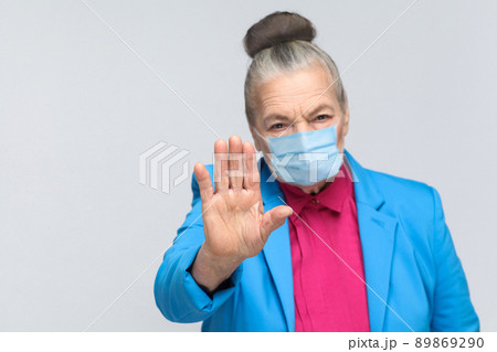 aged grandma with surgical medical mask showing stop sign at camera. Grandmother with light blue suit and pink shirt standing with collected bun hair. indoor studio shot, isolated on gray background 89869290
