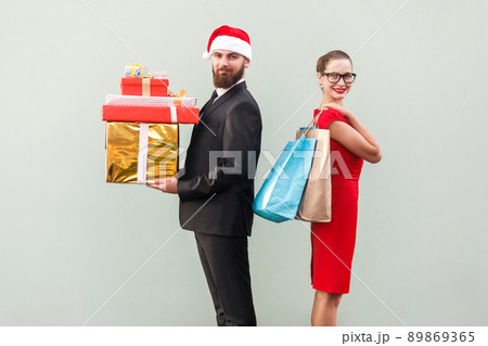 Back to back. Profile view man in red hat and woman in red dress and glasses. Happiness couple holding christmas gifts and colorful packages and looking at camera with toothy smiling. Studio shot Back to back. Profile view man in red hat and woman in red dress and glasses. Happiness couple holding christmas gifts and colorful packages and looking at camera with toothy smiling. Studio shot 89869365