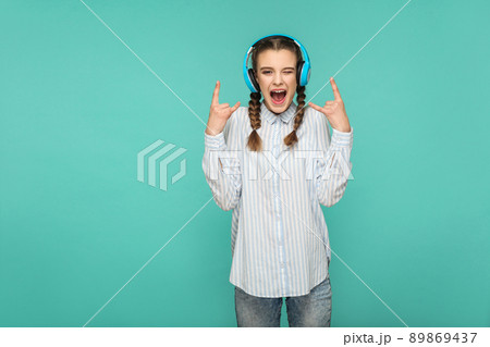 happy amazed girl in striped blue shirt standing listening favorite music with headphone, looking at camera with surprised face and rock sign, Indoor studio shot, isolated on blue or green background 89869437