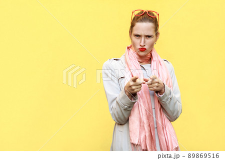 The serious woman, wearing coat, having anger looks, pointing finger at camera. Isolated shot on yellow wall The serious woman, wearing coat, having anger looks, pointing finger at camera. Isolated shot on yellow wall 89869516