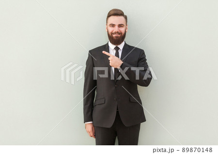 Young handsome bearded businessman with beaming smile pointing with finger at capy space and looking at camera. Indoor shot, gray background 89870148