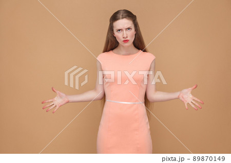 unhappy anger woman with hands up looking at camera. emotional cute, beautiful woman with makeup and long hair in pink dress. indoor, studio shot, isolated on light brown or beige background. 89870149