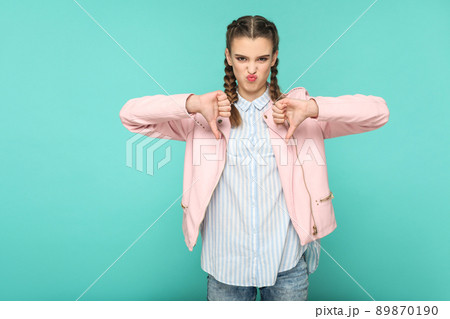 unsatisfied dislike portrait of beautiful cute girl standing with makeup and brown pigtail hairstyle in striped light blue shirt pink jacket. indoor, studio shot isolated on blue or green background. 89870190