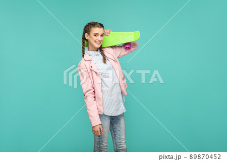 happy beautiful girl in casual style, pigtail hair and pink jacket, standing and holding skateboard and looking at camera with toothy smile, Indoor studio shot, isolated on blue or green background happy beautiful girl in casual style, pigtail hair and pink jacket, standing and holding skateboard and looking at camera with toothy smile, Indoor studio shot, isolated on blue or green background 89870452