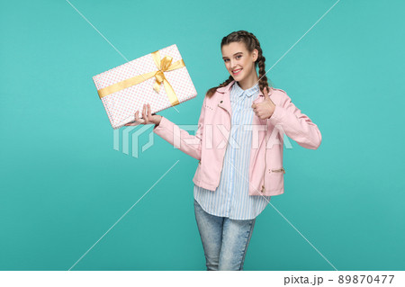 happy thumbs up beautiful girl in casual style and pink jacket, standing and holding dotted gift box and looking at camera with satisfied face, Indoor, isolated on blue or green background 89870477
