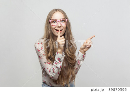 Happiness beautiful young girl, showing finger at copy space and looking at camera with conspiracy sign. Studio shot, isolated on gray background 89870596