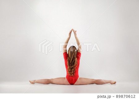 young athletic blonde woman in red leotard sitting on string. hanumanasana yoga pose. isolated indoor studio shot on light gray background. healthy lifestyle and leisure activity concept. 89870612