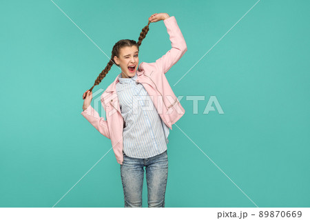 portrait of happy beautiful funny girl with blue striped t-shirt and pink jacket standing looking at camera, holding her pigtail hair and winking. indoor studio shot isolated on green background. portrait of happy beautiful funny girl with blue striped t-shirt and pink jacket standing looking at camera, holding her pigtail hair and winking. indoor studio shot isolated on green background. 89870669