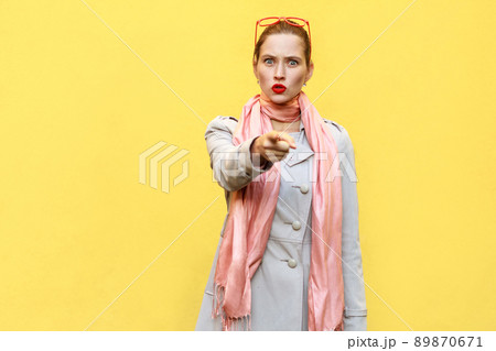 The beautiful woman, wearing autumn coat, opening mouths widely, having surprised shocked looks, pointing finger at camera. Studio shot, yellow wall The beautiful woman, wearing autumn coat, opening mouths widely, having surprised shocked looks, pointing finger at camera. Studio shot, yellow wall 89870671