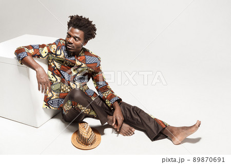 Wondering afro man in traditional national clothes sit on cube. Indoor, isolated on gray background 89870691