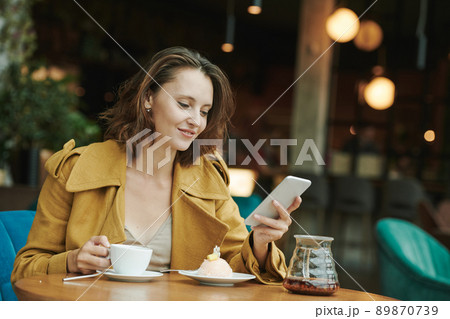 Young bearded manager sitting at table with coffee cup and croissant and recording voice message for colleague using smartphone in cafe Young bearded manager sitting at table with coffee cup and croissant and recording voice message for colleague using smartphone in cafe 89870739