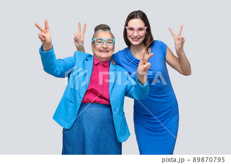 Grandmother with Granddaughter showing peace or victory sign, looking at camera with toothy smile. Relations in the family. happiness and family relationship. Studio shot, isolated on gray background 89870795