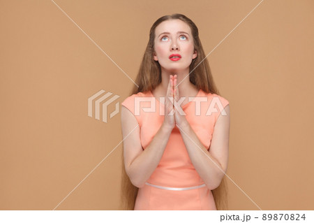 Oh God please forgive me. portrait of emotional cute, beautiful woman with makeup and long hair in pink dress. indoor, studio shot, isolated on light brown or beige background. 89870824