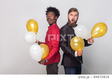 Two shocked friend wearing red shirt and black jacket holding many air balloon and looking at camera with shocked face. Indoor shot 89870828