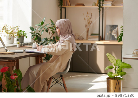 Young Muslim woman wearing headscarf sitting alone in cozy office room surfing Internet on smartphone during break, copy space 89871330