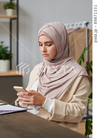 Vertical medium portrait of modern Muslim woman with serious facial expression texting message on her smartphone Vertical medium portrait of modern Muslim woman with serious facial expression texting message on her smartphone 89871331