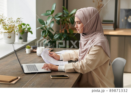 Serious young woman wearing hijab sitting at desk working on new business project alone in modern office 89871332