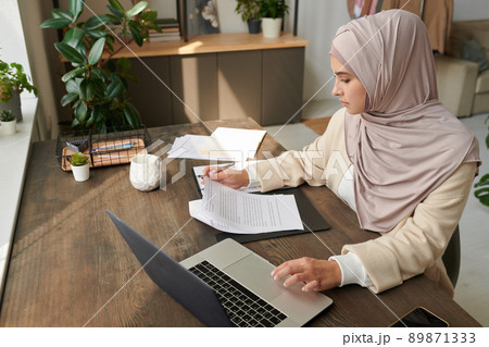 Horizontal high angle view shot of young Middle Eastern woman in hijab working with financial documents in office room Horizontal high angle view shot of young Middle Eastern woman in hijab working with financial documents in office room 89871333