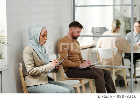 Caucasian man and Muslim woman in hijab sitting on chairs in embassy or visa service agency filling in application forms while waiting in queue 89873316