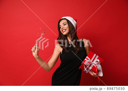 Beautiful woman wearing Santa hat with Christmas gift and bengal lights on red background 89875030