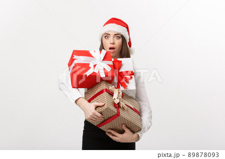 Business Concept - Beautiful young caucasian business woman with santa hat holding a lot of gift boxes with surprising facial expression. 89878093